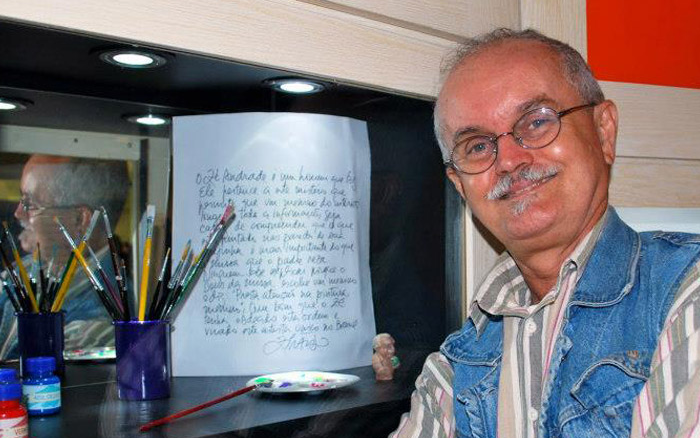 Descrição da foto: homem sorridente posa para foto ao lado de um nicho espelhado na parede onde estão dispostos pincéis, lápis e canetas. 