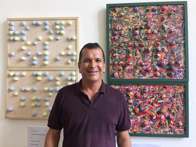 O artista plástico Adel Gonzaga e dois quadros de sua autoria, ambos colagens multicoloridas de objetos redondos sobre a tela formando nomes de alunos dele escritos em braille.