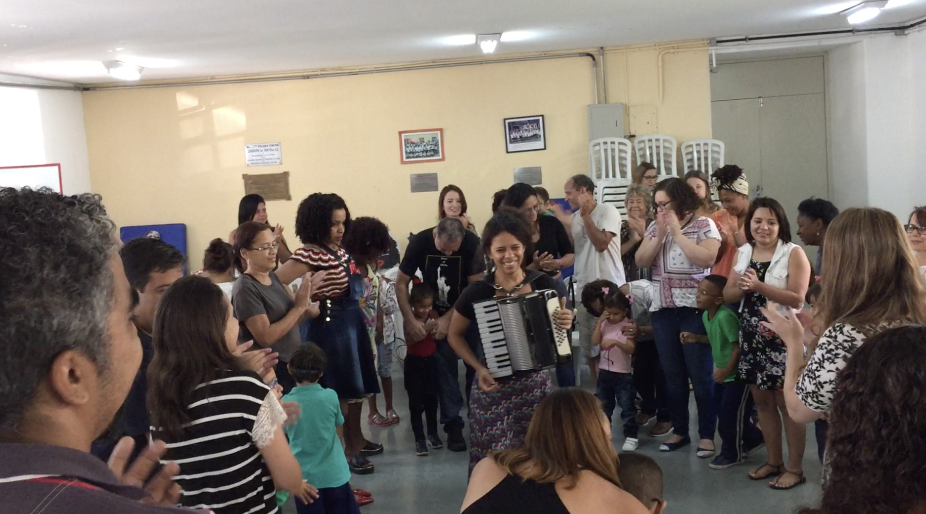 descrição da foto: pais, alunos e professores, cantam e dançam de mãos dadas em roda.  Ao centro, uma mulher toca acordeon.