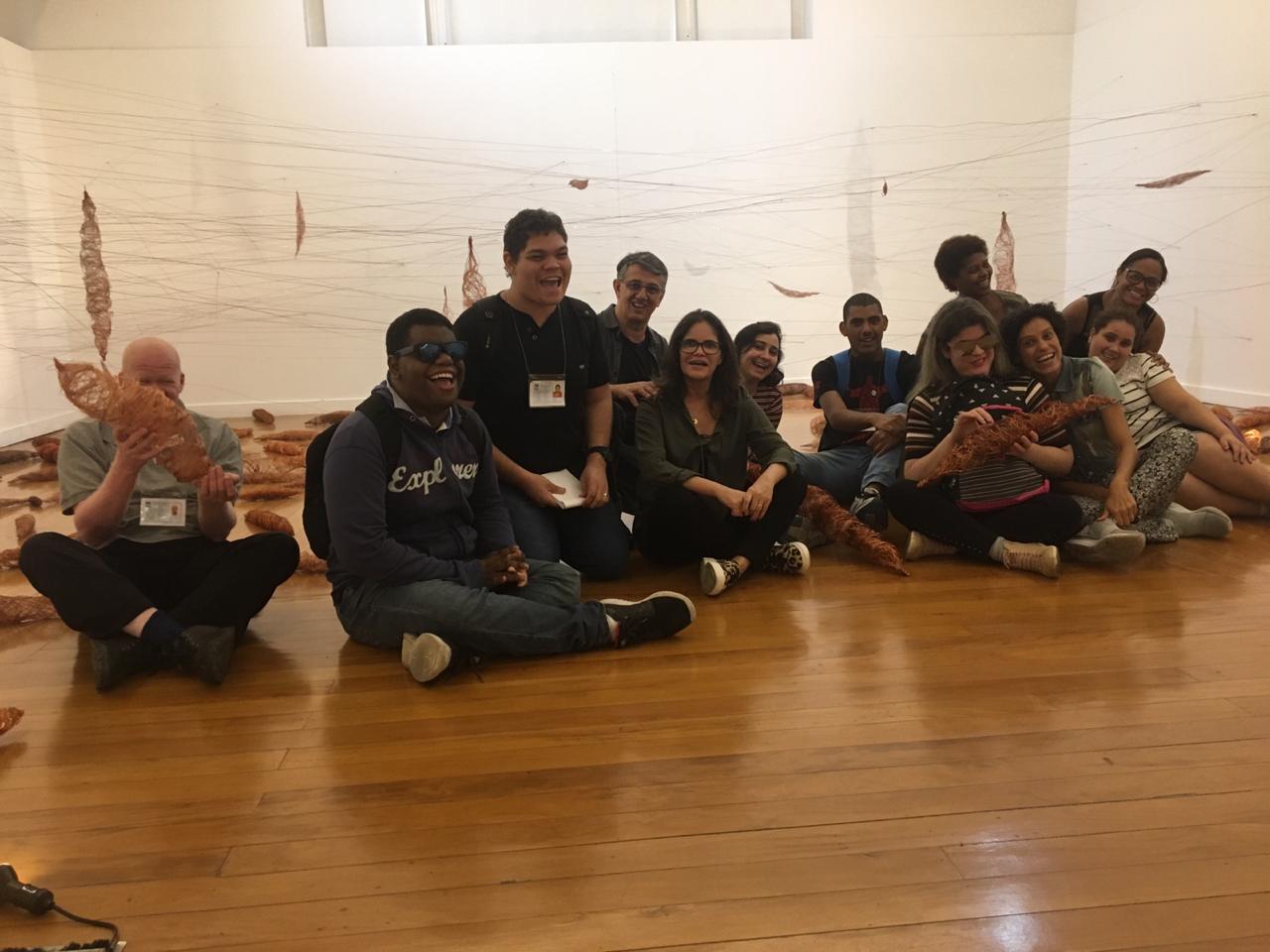 Alunos e professores do IBC sentados no chão de tábua corrida, tendo ao fundo uma instalação artística