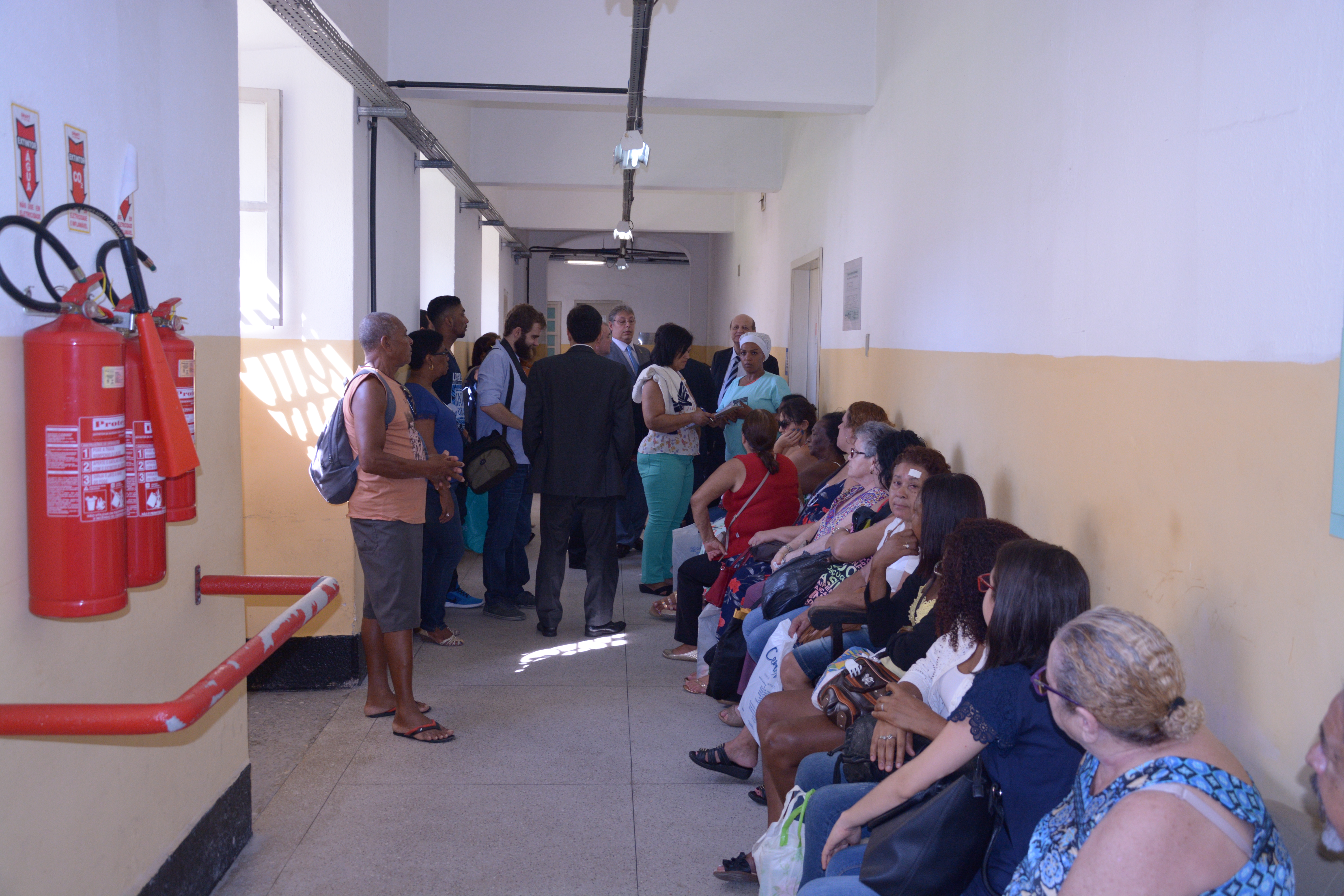 Descrição da foto: corredor com bancos encostados na parede ocupados por pessoas à espera de atendimento médico