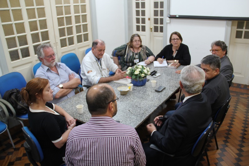 Descrição da foto: sala de reunião com mesa oval de granito ao redor da qual estão sentados seis homens e três mulheres