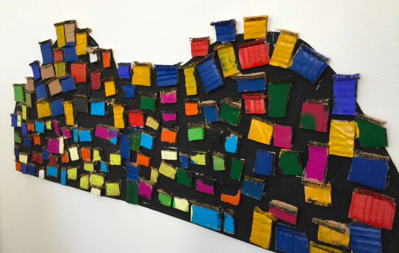 Descrição da foto: painel formado por pedaços de borracha multicoloridos formando um mosaico representando casinhas de uma favela no morro.