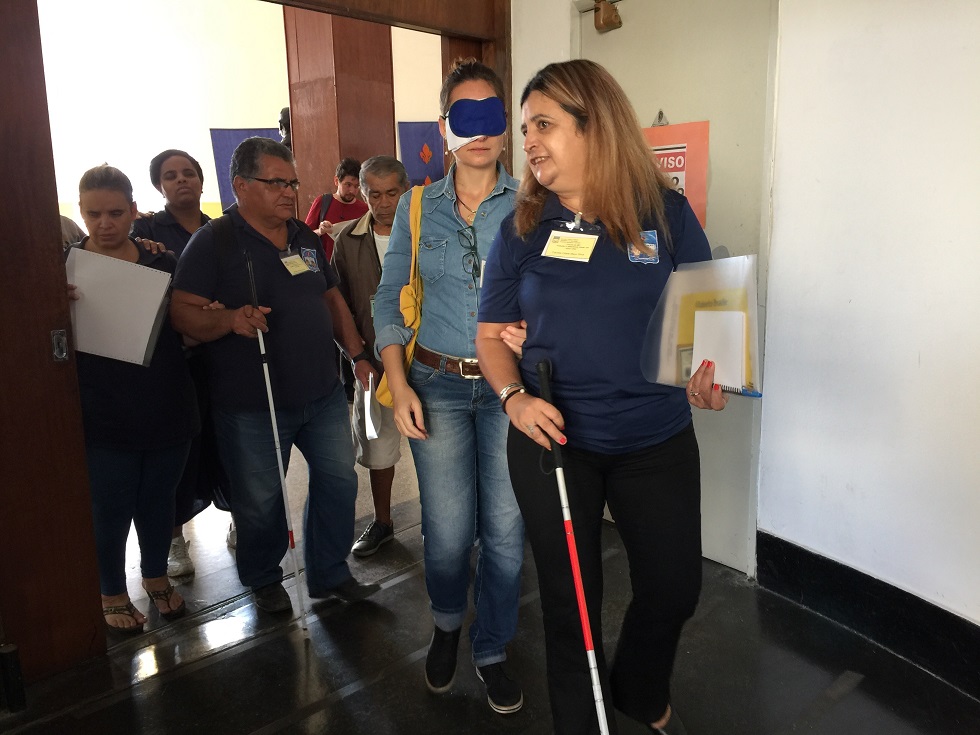 Descrição da foto: mulher vendada sendo guiada por outra mulher cega, com bengala, tendo atrás de si outras pessoas cegas e videntes entrando em um ambiente.