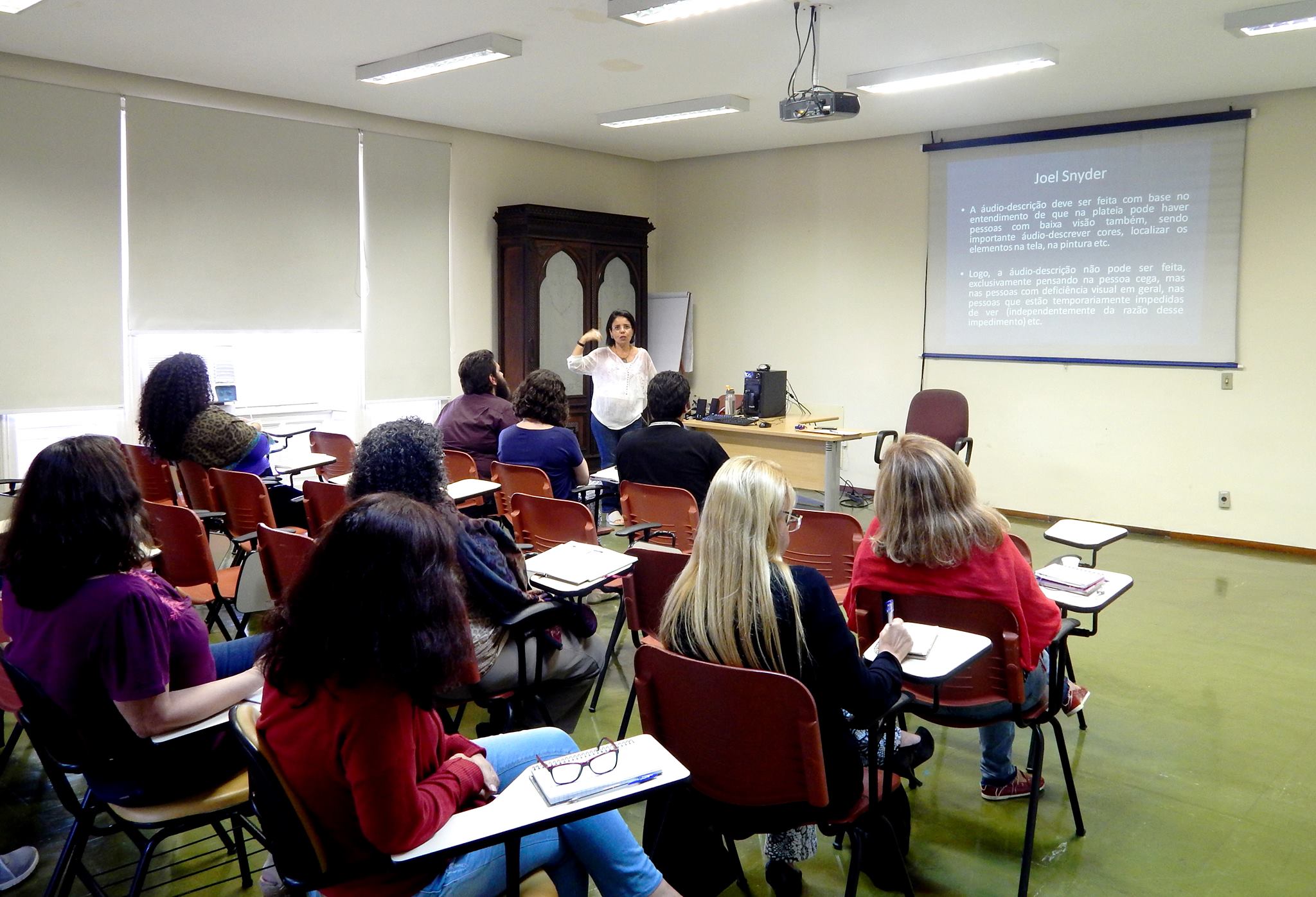 Na sala de aula do MHN, com um armário de madeira no canto esquerdo, Ana Fátima, em pé, ao lado da mesa, está à frente dos cursistas sentados em cadeiras universitárias, no telão, há a projeção de um texto