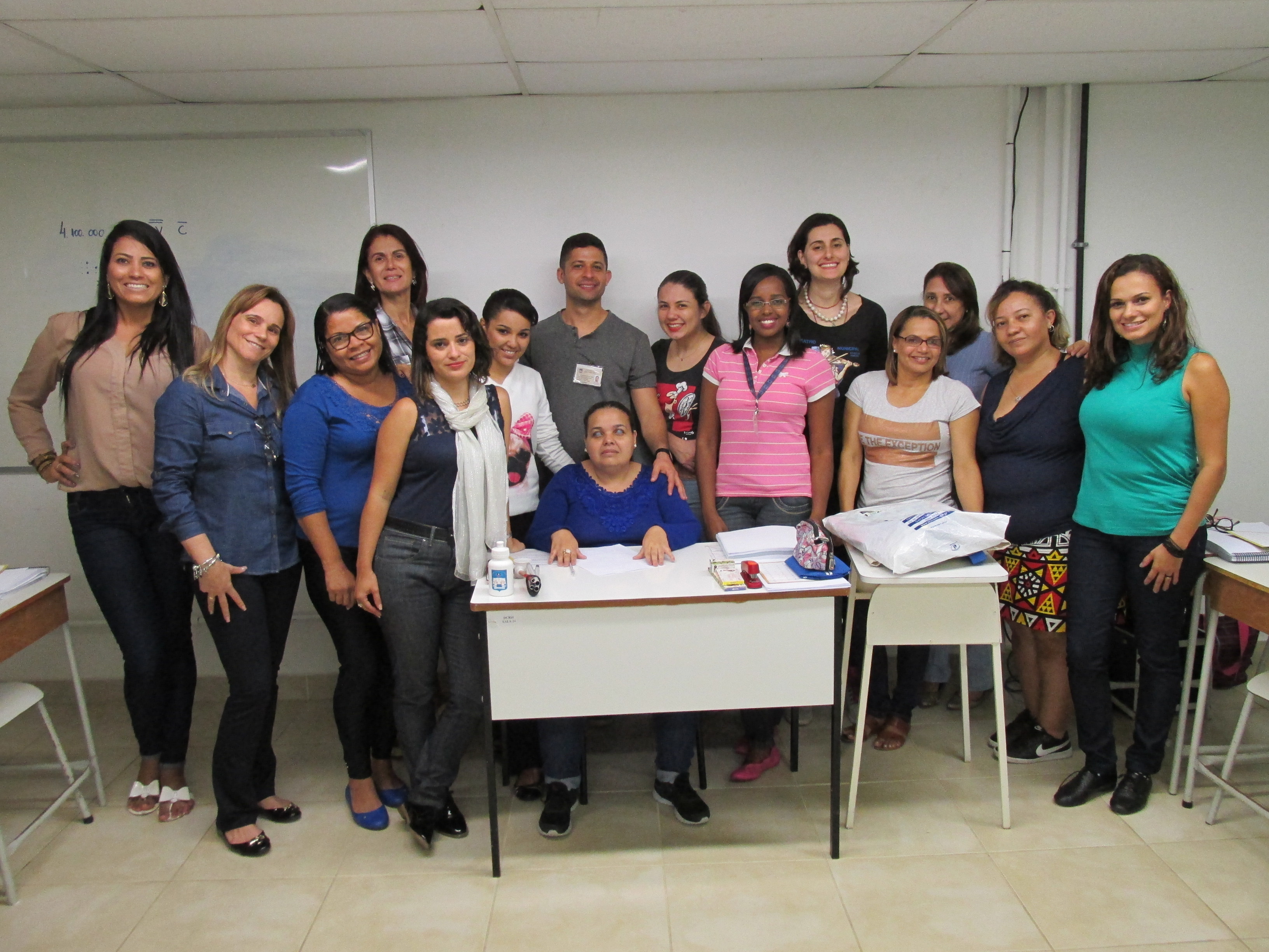 Descrição da imagem: sala de aula. Professora Geni de Abreu posa para a foto sentada à mesa, tendo 13 mulheres e um homem atrás de si.