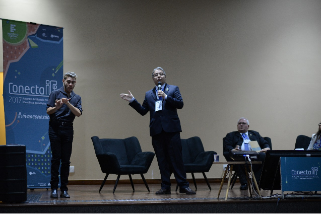 Descrição da foto: no centro de um palco, um homem discursa; ao lado esquerda, um rapaz faz a tradução do discurso para Libras.