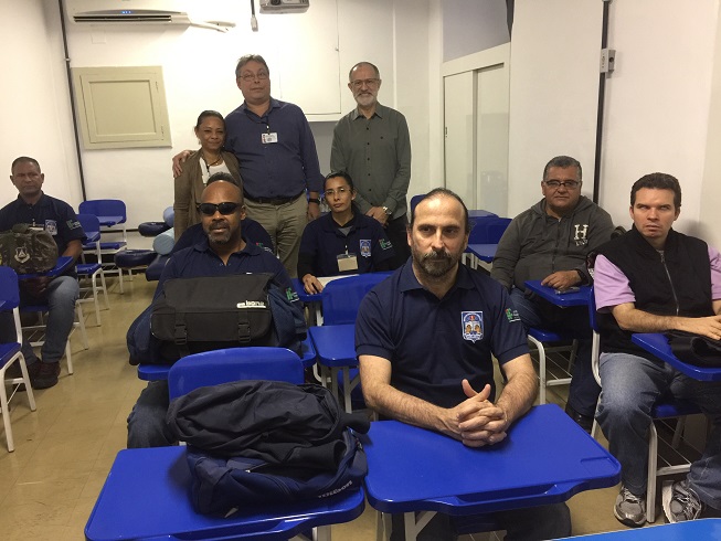 Descrição da foto: sala de aula, onde aparecem, de frente,seis alunos adultos, do sexo masculino e uma aluna do sexo feminino, com camisetas azuis de uniforme.  Ao fundo, dois homens e uma mulher de pé. 