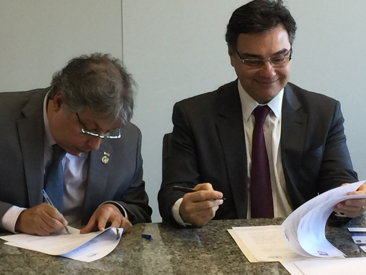 Descrição da foto: dois homens, vestidos de terno e gravata, assinam um documento.