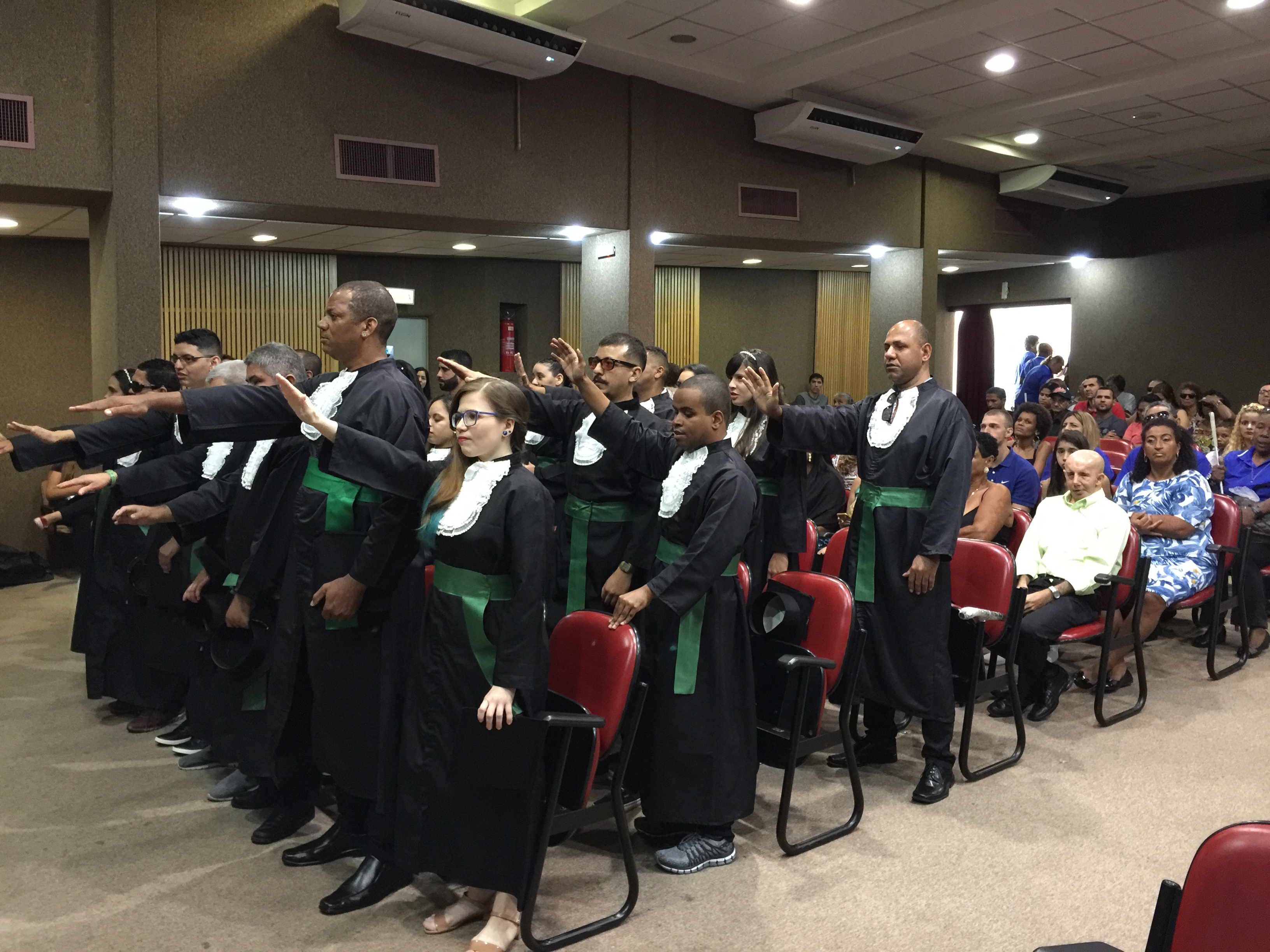 Homens e mulheres vestidos de beca preta, com faixa verde, com o braço direito erguido em posição de juramento.