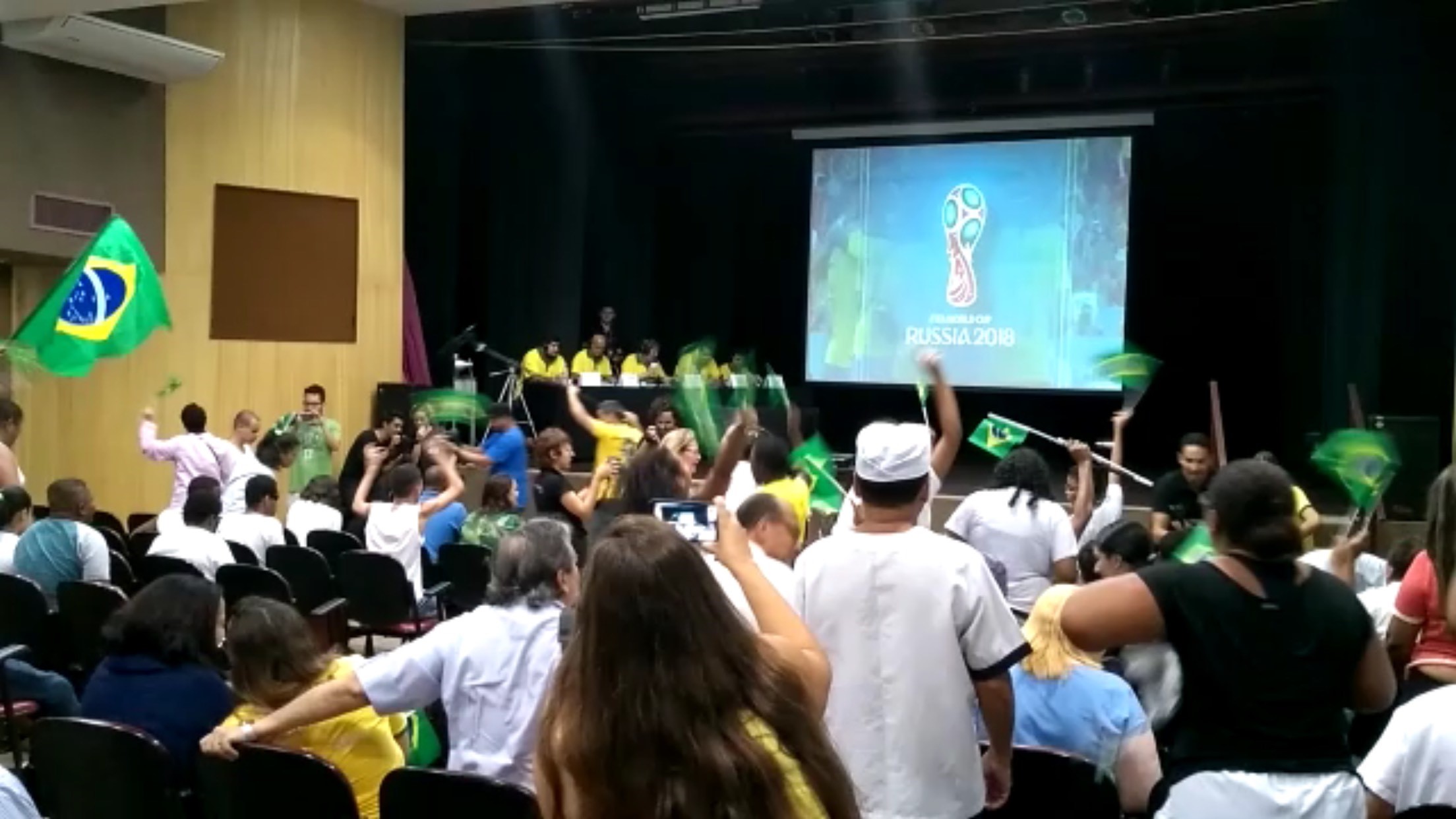 Plateia do Teatro Benjamin Constant bastante cheia de crianças e adultos de frente para o palco, com bandeirinhas do Brasil nas mãos.  No palco, cinco homens de camisetas amarelas, sentados lado a lado a uma mesa retangular virada para o público. Do lado esquerdo deles, um telão em que aparece imagem de uma partida de futebol.