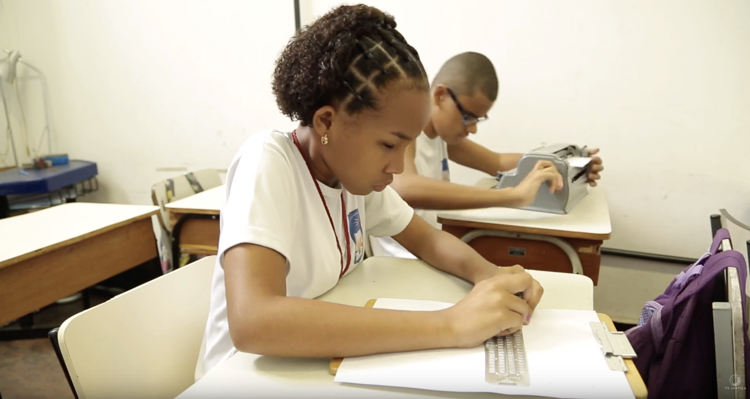 Sala de aula com dois alunos: uma menina em primeiro plano, escrevendo em braille com reglete, e um menino ao fundo, datilografando em uma máquina Perkins.