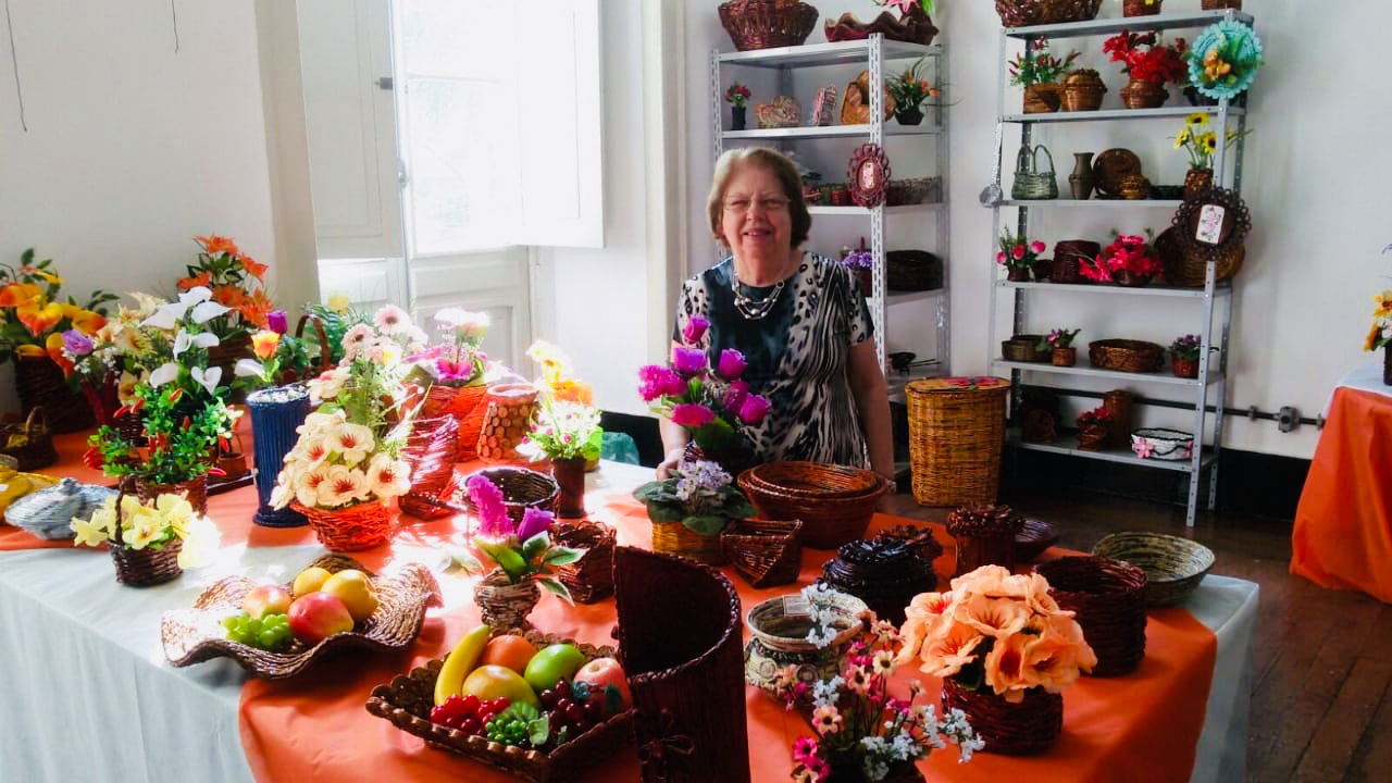Descrição da foto: senhora idosa de pé, por trás de uma mesa repleta de arranjos florais.