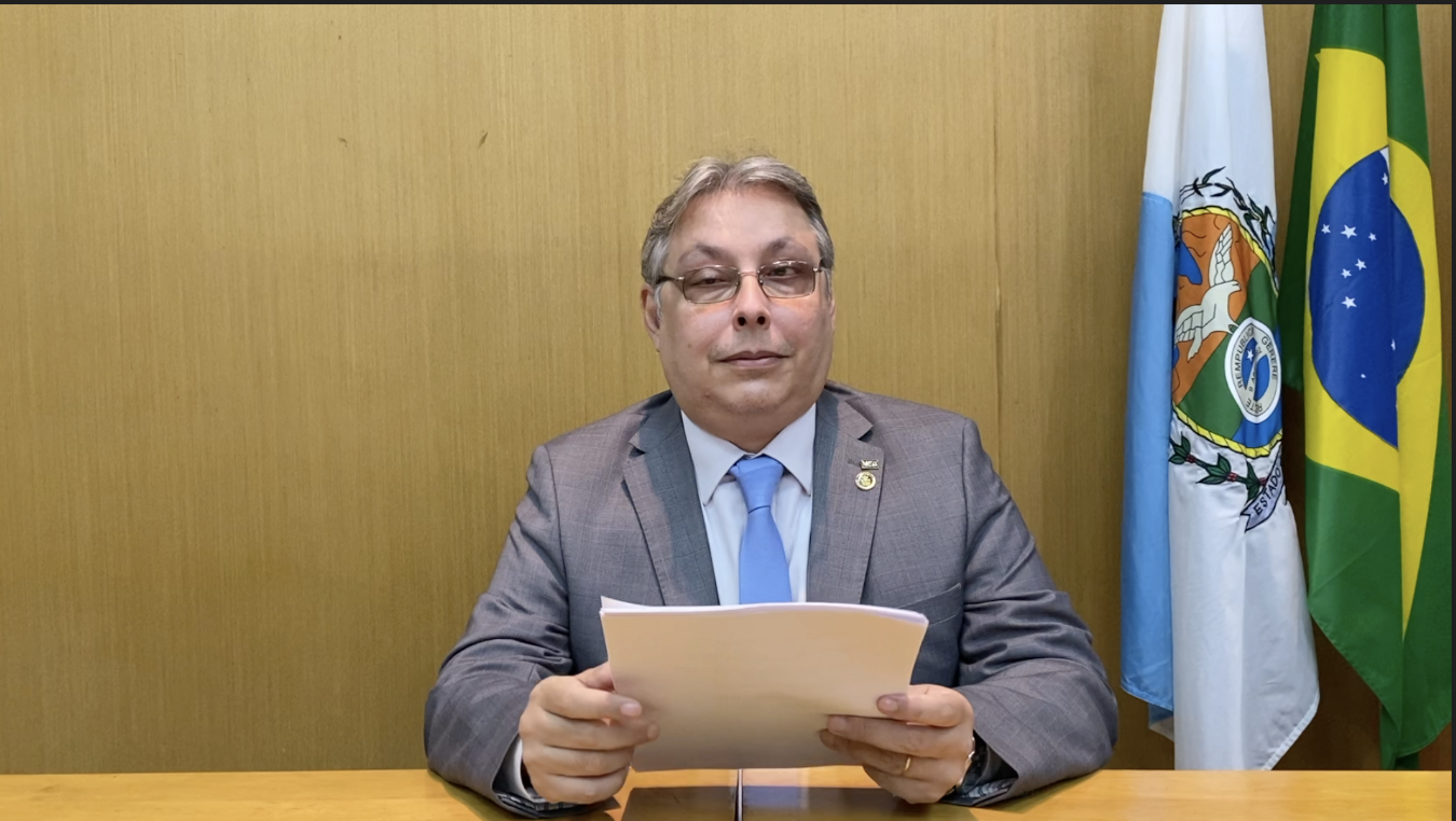 No centro da tela, sentado por trás de uma bancada de madeira, o diretor-geral do IBC, com terno cinza, camisa branca e gravata azul clara.  Do lado direito da tela, as bandeiras do Brasil e do Estado do Rio de Janeiro.